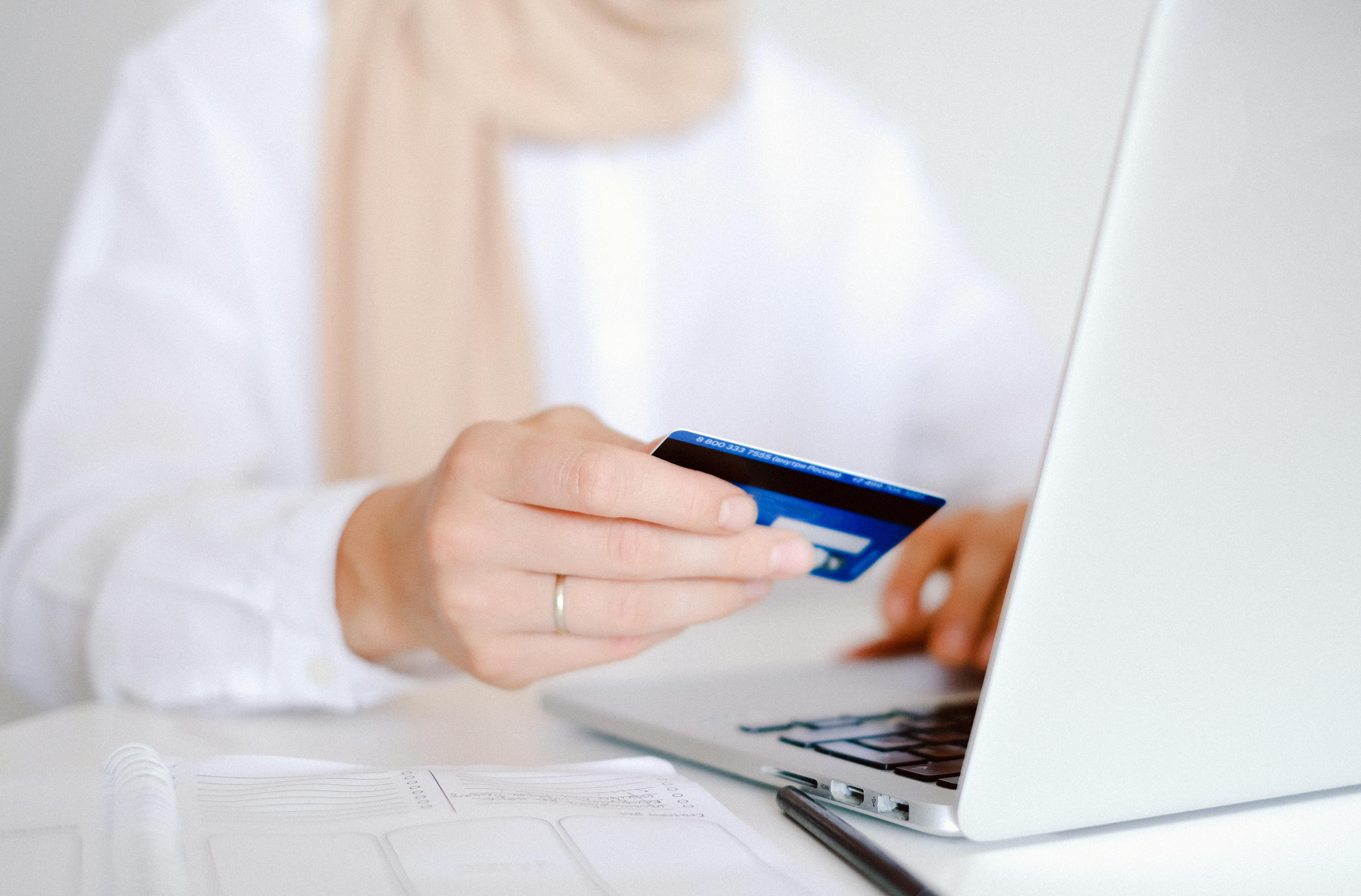 Person holding a blue credit card while using a laptop at a bright desk with a notebook and pen nearby